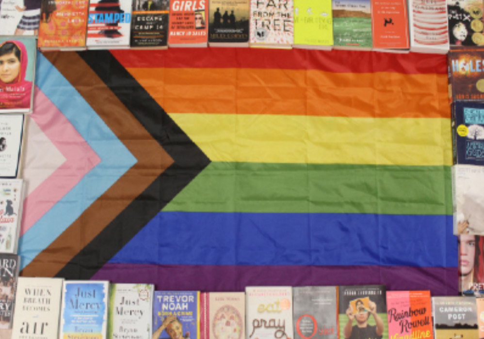 An LGBTQ+ flag surrounded by books, many of which are banned in some states. New laws for California have recently been signed to keep these books in public libraries.