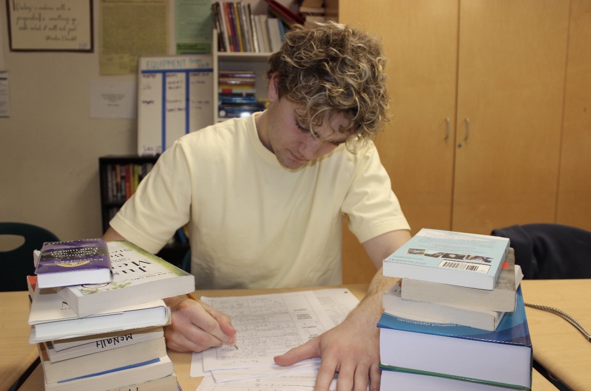 A student finishes math homework with an overload of books surrounding him. He completes
each task, highlighting homework overload and student burnout.